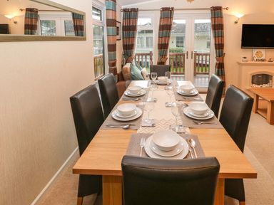 A dining area with a wooden table set for six people with black chairs and glass doors with curtains at 16 Heritage Park in Heritage Park near Kilgetty