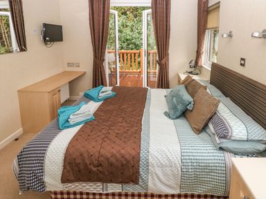 A bedroom with a bed covered by patterned bedding and pillows a desk a mounted tv and glass doors opening to a balcony at 16 Heritage Park Heritage Park near Kilgetty