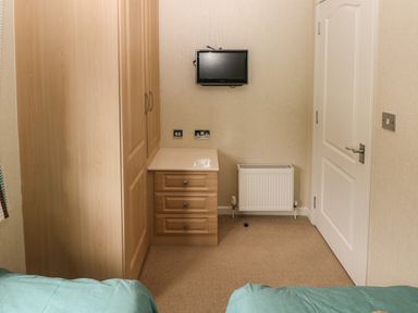 A small bedroom with a wooden wardrobe and chest of drawers a wall-mounted TV a radiator and a closed white door at 16 Heritage Park in Heritage Park near Kilgetty