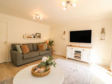 A living room with a gray sofa cushions a white coffee table with a wicker tray and white flowers a white media console and wall-mounted TV at Amaranta Brewers Quay Harbour