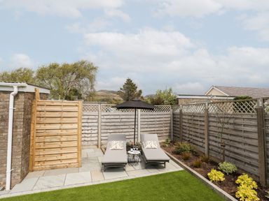 A backyard with two lounge chairs under an umbrella a small table with bottles and glasses and a garden bed along a wooden fence at 31 Beach Road in Morfa Bychan