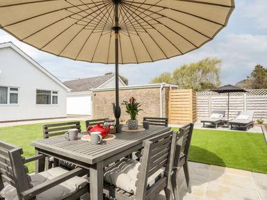 An outdoor patio with a table and chairs under a large umbrella with cups and a teapot on the table and sun loungers in the background at 31 Beach Road in Morfa Bychan