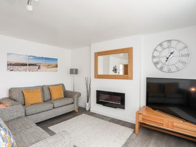 A living room with grey sofas yellow cushions a wooden TV stand and a wall clock at 31 Beach Road Morfa Bychan