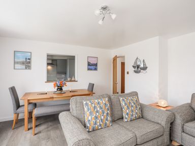 A living room with grey sofas and patterned cushions beside a dining table with chairs and a bench at 31 Beach Road in Morfa Bychan