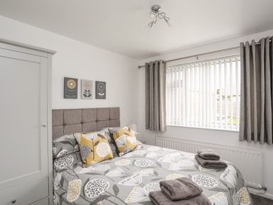 A bedroom with a bed with floral bedding towels on the bed a wardrobe and window with vertical blinds and curtains at 31 Beach Road in Morfa Bychan