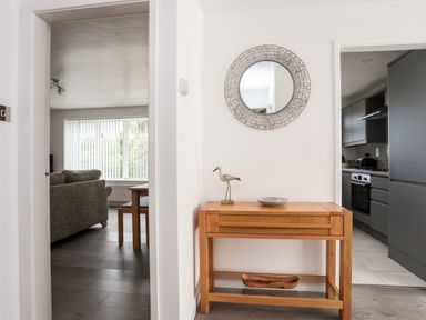A hallway with a wooden console table with a bird sculpture and bowl under a round wall mirror between a living room and a kitchen at 31 Beach Road in Morfa Bychan