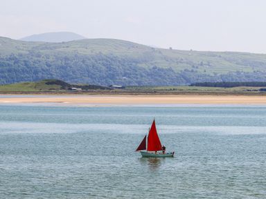 A small sailboat with red sails on a body of water with hills and sandy shore in the background at 31 Beach Road in Morfa Bychan