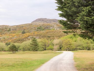 A gravel path leading through a grassy area with trees and hills in the background at 31 Beach Road in Morfa Bychan