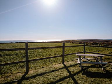A wooden picnic table next to a wooden fence overlooking fields and the sea at Faraway Cottage in St Levan near Porthgwarra