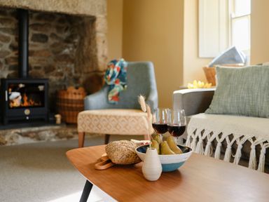 A living room with a coffee table holding bread pears and wine glasses by a stone fireplace at Faraway Cottage in St Levan near Porthgwarra