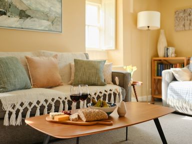 A living room with a sofa, pillows, a wooden coffee table with bread, cheese, two glasses of red wine, pears, and a small vase at Faraway Cottage in St Levan near Porthgwarra