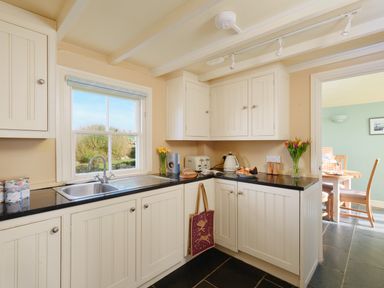 A kitchen with white cabinets black countertops a stainless steel sink a window and flowers on the counter at Faraway Cottage in St Levan near Porthgwarra