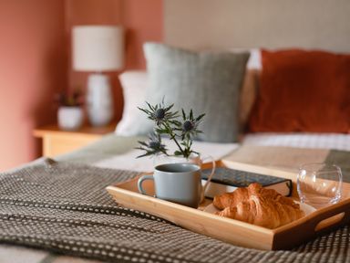 A tray with croissants a cup a glass and a book on a bed with cushions at Faraway Cottage in St Levan near Porthgwarra