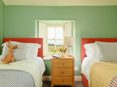A bedroom with two single beds with red headboards and a wooden bedside table with a lamp and flowers near a window at Faraway Cottage in St Levan near Porthgwarra