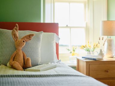 A bed with a stuffed bunny and book beside pillow next to a wooden nightstand with lamp and flowers at Faraway Cottage in St Levan near Porthgwarra