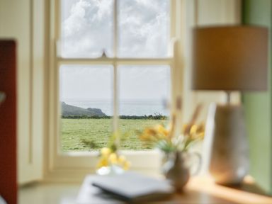 A view through a window showing a green field hills and ocean with a lamp a vase with flowers and a book inside a room at Faraway Cottage in St  Levan near Porthgwarra