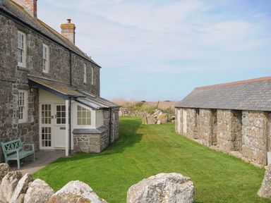 A stone building with a white door and windows next to a stone wall and grassy yard at Three Chimneys in St Levan near Porthgwarra