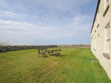 A grassy yard with a wooden table and benches surrounded by a stone wall and a building at Three Chimneys in St Levan near Porthgwarra