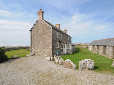 A stone house with chimneys next to a stone building and a grassy area with rocks at Three Chimneys in St Levan near Porthgwarra
