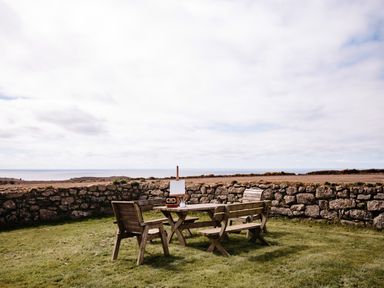 An outdoor wooden table with chairs and an easel on grass next to a stone wall with a field and ocean in the background at Three Chimneys in St Levan near Porthgwarra