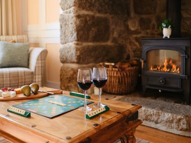 A living room with a Scrabble game and two glasses of wine on a wooden table near a lit fireplace at Three Chimneys in St Levan near Porthgwarra