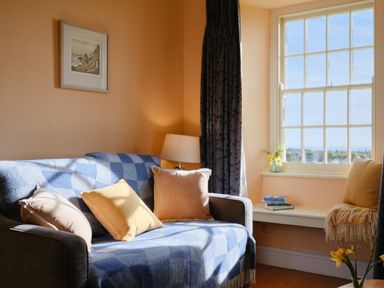 A room with a gray sofa covered with a blue checkered blanket and cushions beside a window with books and flowers at Three Chimneys in St Levan near Porthgwarra