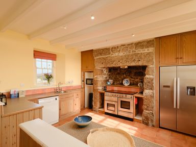 A kitchen with wooden cabinets a stone fireplace a stainless steel refrigerator and a window with flowers at Three Chimneys in St Levan near Porthgwarra