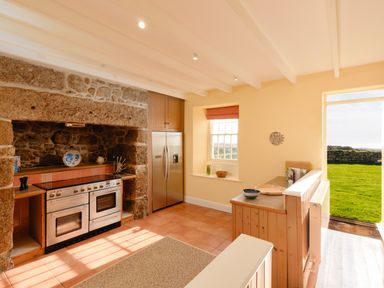 A kitchen with a stone fireplace oven stainless steel refrigerator wooden cabinets and an open door to a grassy yard at Three Chimneys in St Levan near Porthgwarra