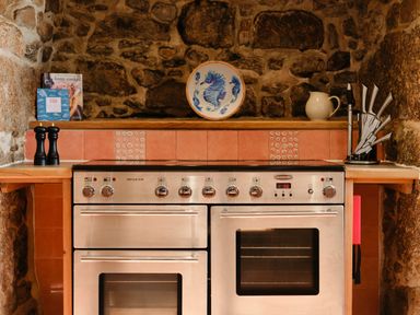 A kitchen oven with stone walls and tiled backsplash with cookbooks and kitchen utensils at Three Chimneys in St Levan near Porthgwarra