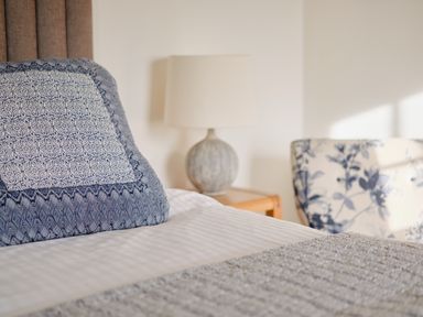 A bed with a blue patterned pillow a lamp on a wooden side table and a floral patterned chair at Three Chimneys in St Levan near Porthgwarra
