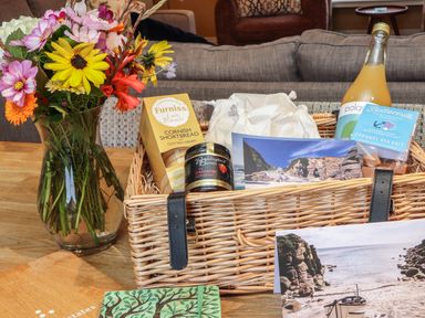 A wicker basket with Cornish shortbread, strawberry conserve, caramel sea salt fudge, a bottle of drink, postcards and a vase of flowers on a wooden table at Higher Roskestal in St Levan near Porthgwarra