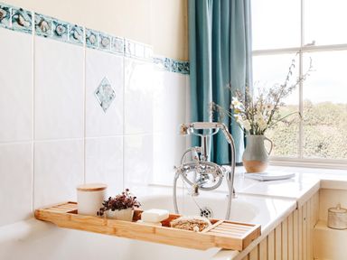 A bathroom with a bathtub and a wooden tray holding toiletries next to a window with blue curtains at Higher Roskestal in St Levan near Porthgwarra