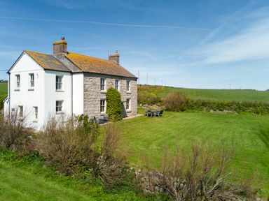 A stone and white painted house with two chimneys and garden furniture on a lawn at Higher Roskestal in St Levan near Porthgwarra