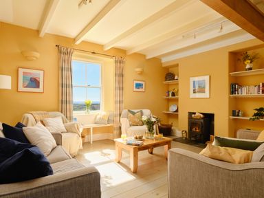 A living room with sofas a wooden coffee table flowers on the table a window with curtains and a fireplace at Higher Roskestal in St Levan near Porthgwarra