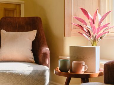 A side table with two mugs and a book next to two chairs and a pink plant at Higher Roskestal in St Levan near Porthgwarra