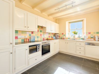 A kitchen with white cabinets colorful tiled backsplash a microwave oven built-in oven a window with flowers on the sill at Higher Roskestal in St Levan near Porthgwarra