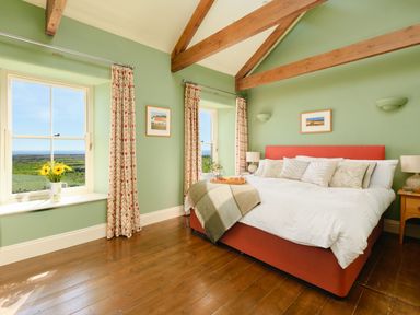 A bedroom with a large bed two windows with curtains and wooden beams on the ceiling at Higher Roskestal in St Levan near Porthgwarra