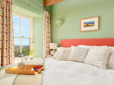 A bedroom with a bed and pillows next to a window with floral curtains and a tray with breakfast at Higher Roskestal in St Levan near Porthgwarra