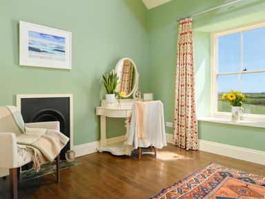 A corner of a room with a chair, a fireplace, a vanity table with a mirror, a window with patterned curtains, and a vase of flowers on the windowsill at Higher Roskestal in St Levan near Porthgwarra