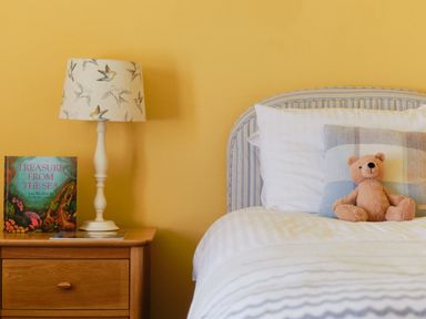 A bedside table with a lamp and a book next to a bed with pillows and a teddy bear at Higher Roskestal in St Levan near Porthgwarra