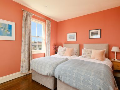 A bedroom with two single beds a window with curtains and framed pictures on coral walls at Higher Roskestal in St Levan near Porthgwarra