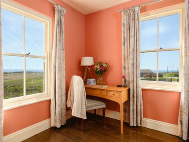 A corner desk with a chair and lamp between two windows in a room with coral walls at Higher Roskestal in St Levan near Porthgwarra