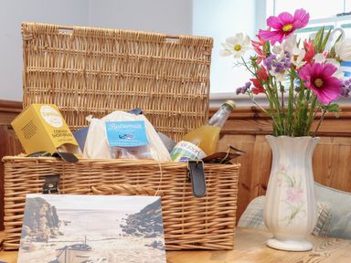 A wicker picnic basket with food items and a bottle next to a floral vase with flowers at Corner Cottage in Porthgwarra