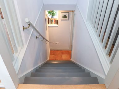 A staircase with grey steps leading down to a small landing with books on a shelf and a framed picture at Corner Cottage in Porthgwarra