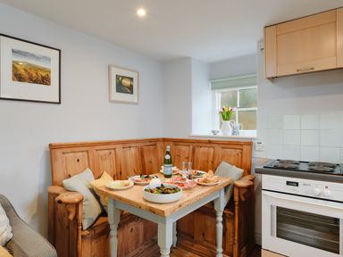 A kitchen dining area with a wooden corner bench and table set with food and drinks at Corner Cottage in Porthgwarra
