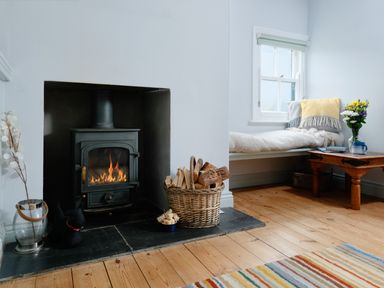 A room with a wood burning stove fire basket with firewood a window seat with cushions and a wooden table with flowers at Corner Cottage in Porthgwarra