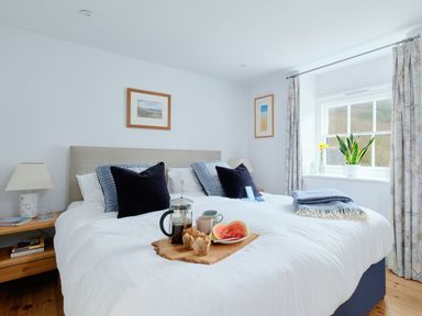A bedroom with a large bed with breakfast on a tray a window with curtains and plants at Corner Cottage in Porthgwarra