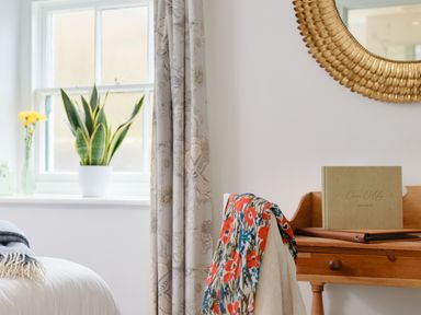 A window with a plant and flowers next to a wooden desk with a book and a chair with a floral cloth at Corner Cottage in Porthgwarra