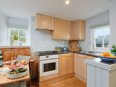 A kitchen with wooden cabinets a stove a wooden table with food and drinks and two windows at Corner Cottage in Porthgwarra