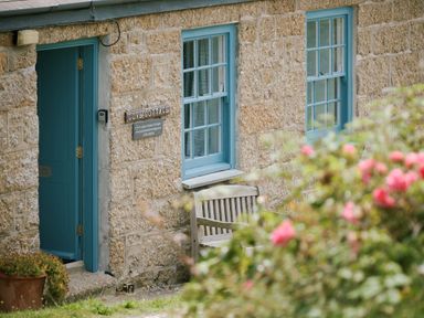 A stone cottage exterior with a blue door and windows a wooden bench and a flower bush at Cove Cottage in Porthgwarra